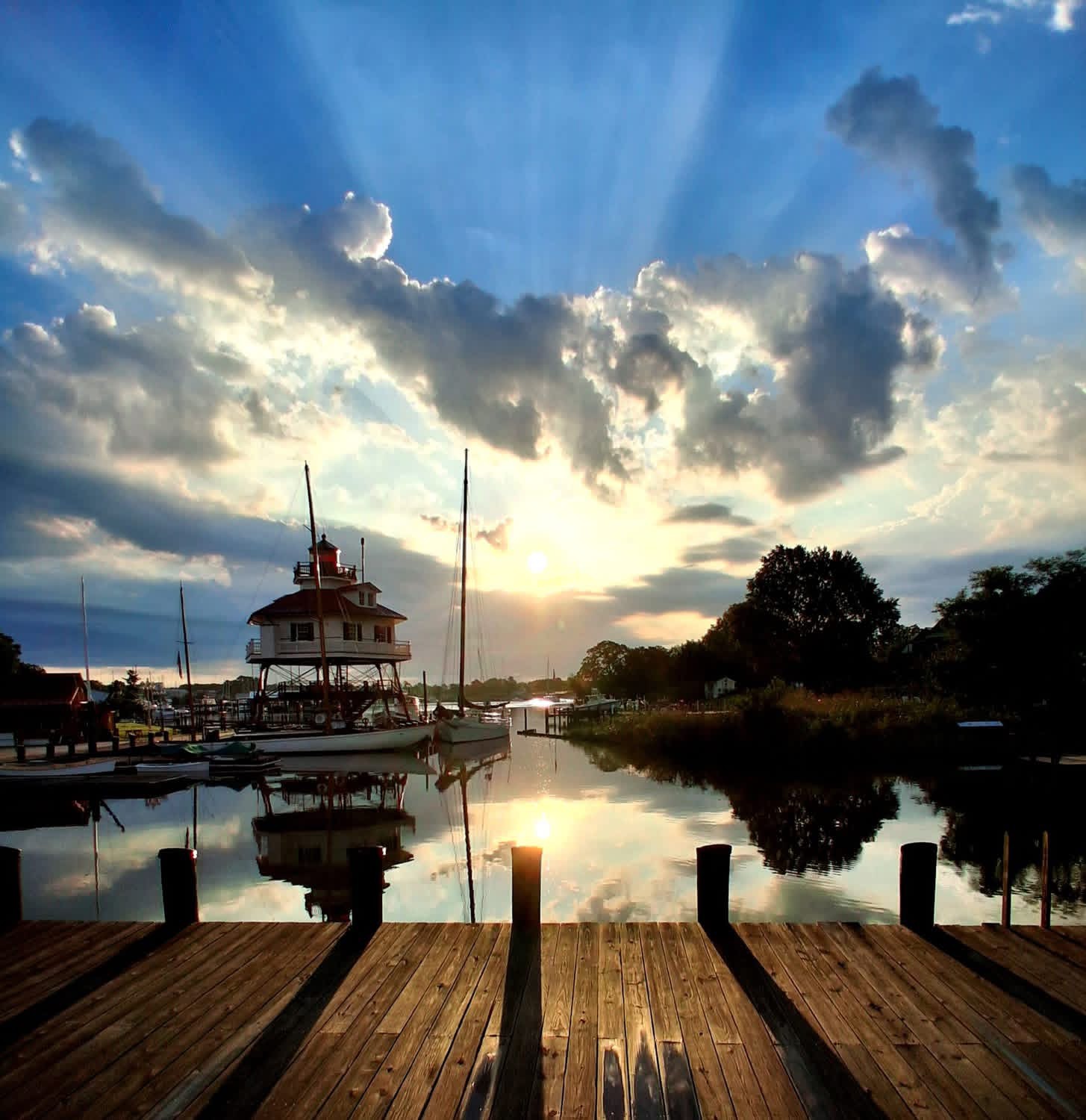 Sunbeams breaking through clouds above a lighthouse and sailboats reflected in a quiet harbor