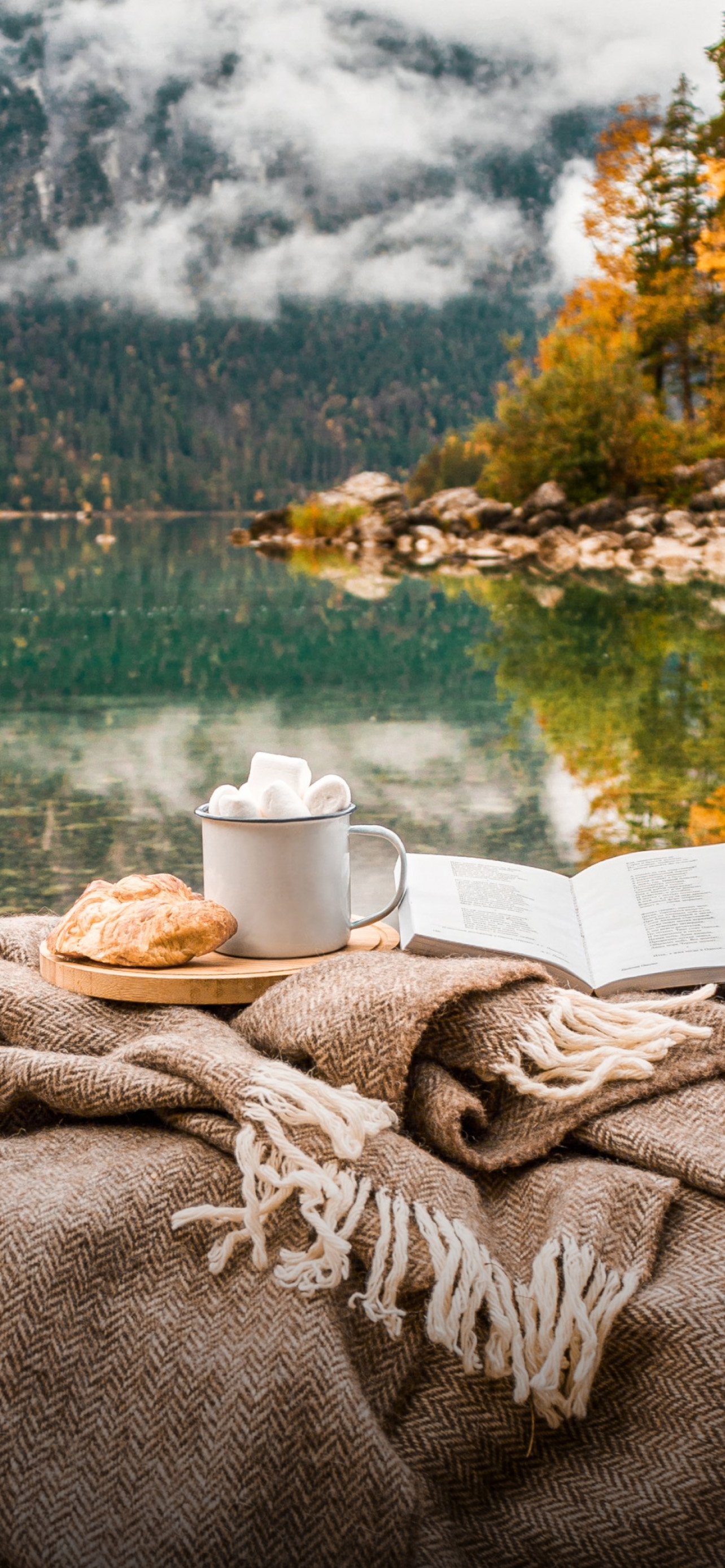 A warm drink, pastry, and open book on a cozy blanket beside an autumn mountain lake